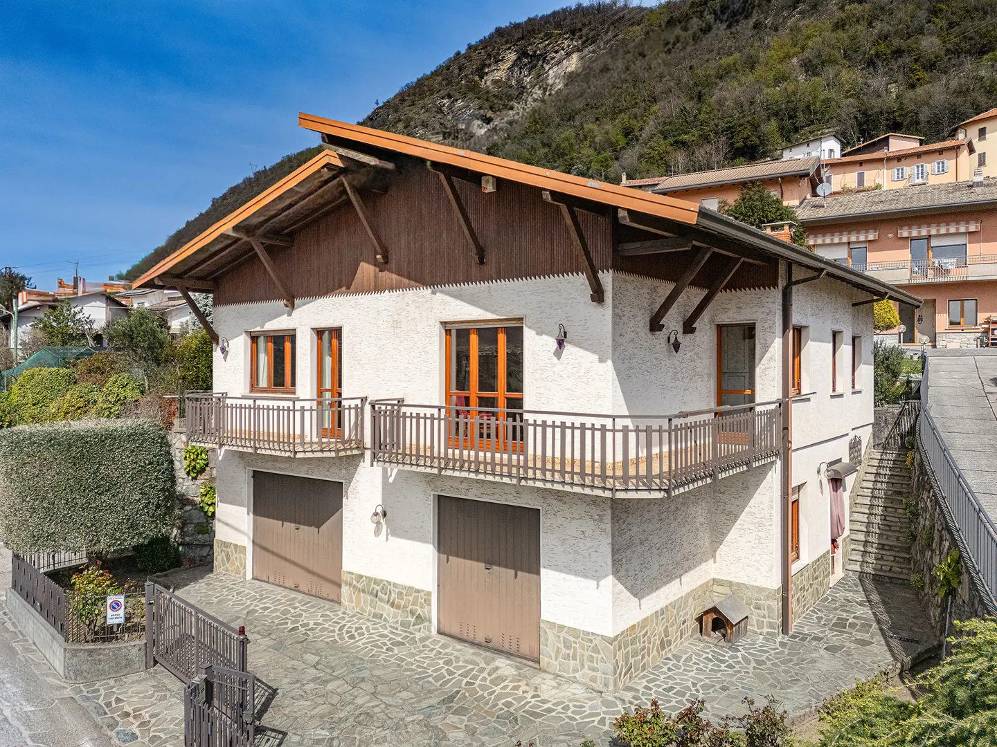Exterior view of a two-story white house with brown trim, two garage doors, and two balconies, set against a mountain backdrop.