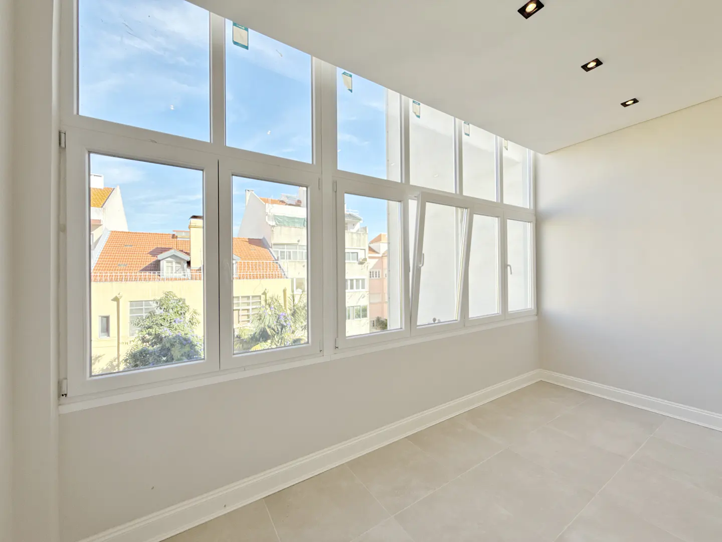 Bright, empty room with large white windows showing a cityscape with red roofs and blue sky.