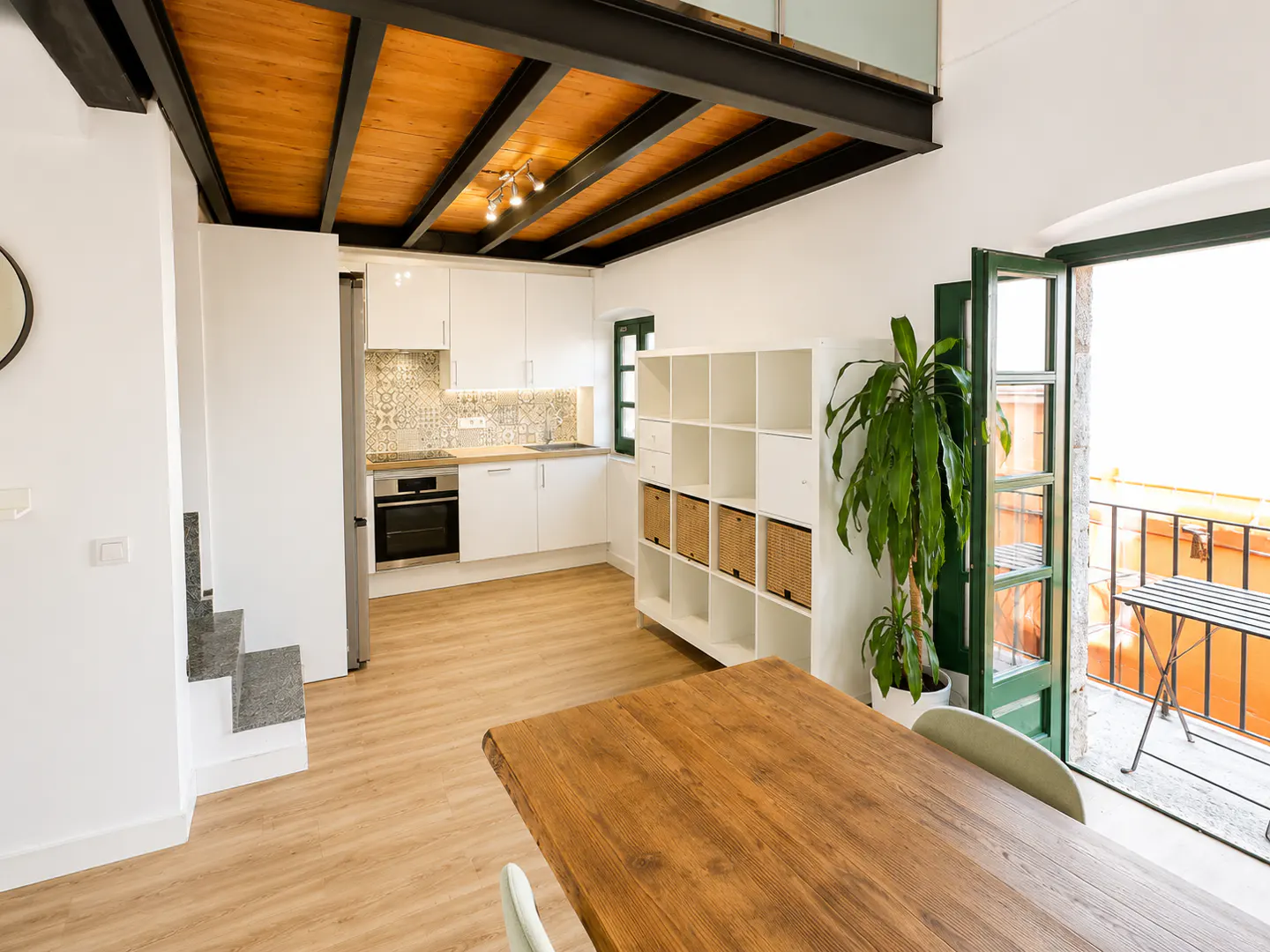 Bright, modern apartment interior with white kitchen, wood table, and open balcony. Black beams support a wood ceiling. A plant sits near the open green door.