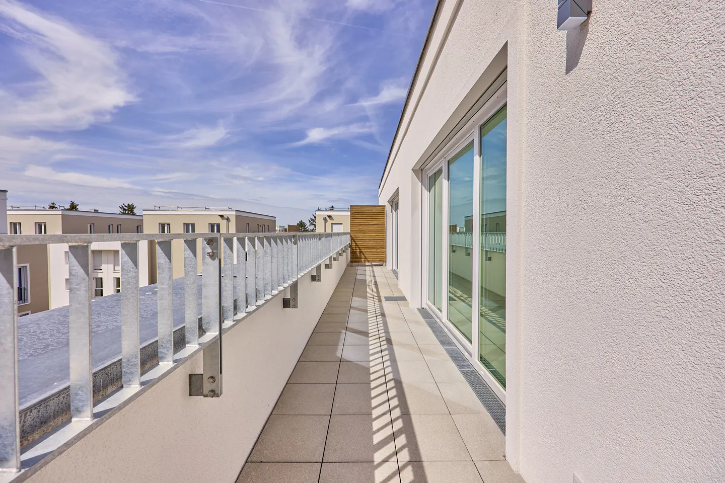 Modern apartment balcony with tiled floor, metal railing, and large glass sliding doors under a bright blue sky.
