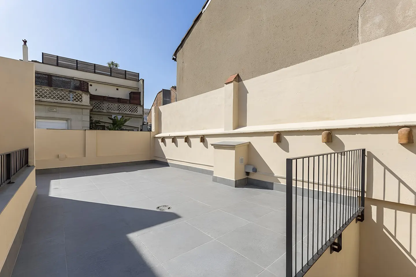 Spacious rooftop terrace with light-colored walls, grey tiled flooring, and black metal railings under a clear blue sky.