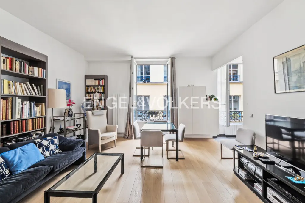 Bright living room with hardwood floors, bookshelves, a blue sofa, and a dining table near a window overlooking a city street.