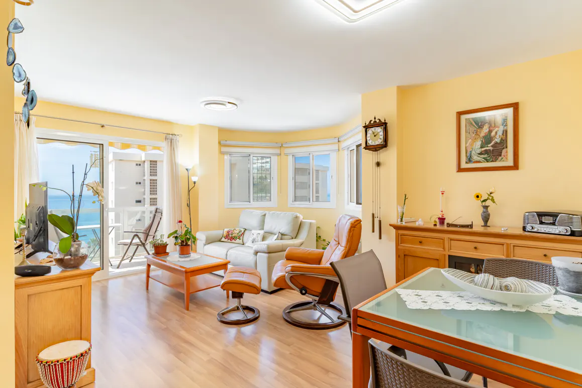 Bright living room with yellow walls, white sofa, orange leather recliner, dining area, and balcony with ocean view.