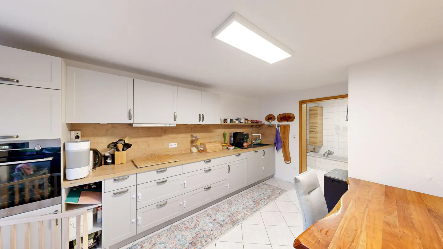 A bright kitchen with white cabinets, light wood countertops, and a patterned rug on white tile floor. A wooden table is in the foreground.