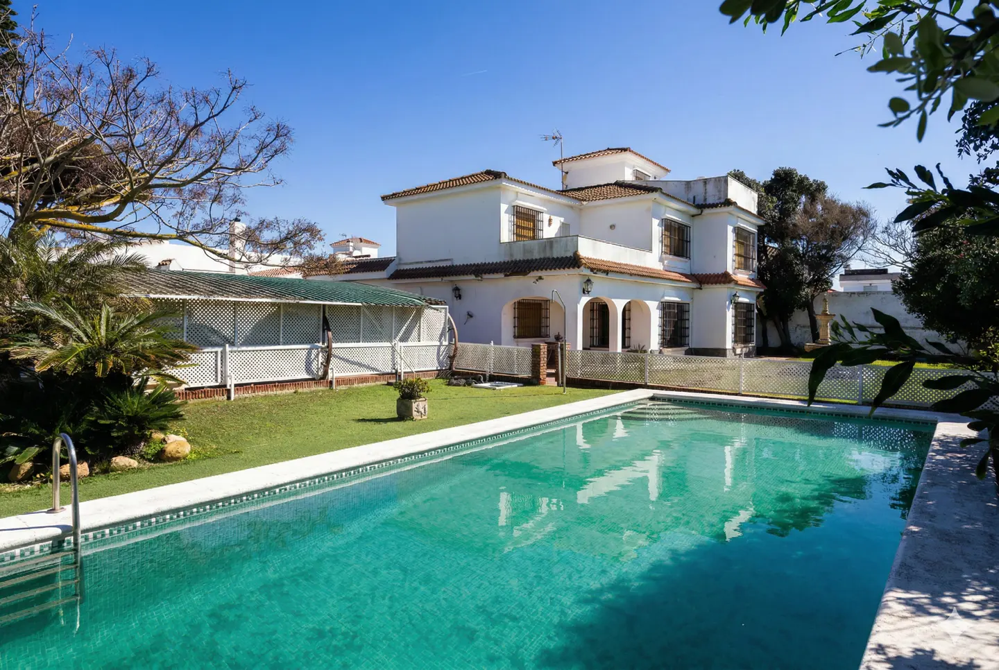 A white two-story house with a red tile roof overlooks a turquoise swimming pool and green lawn on a sunny day.