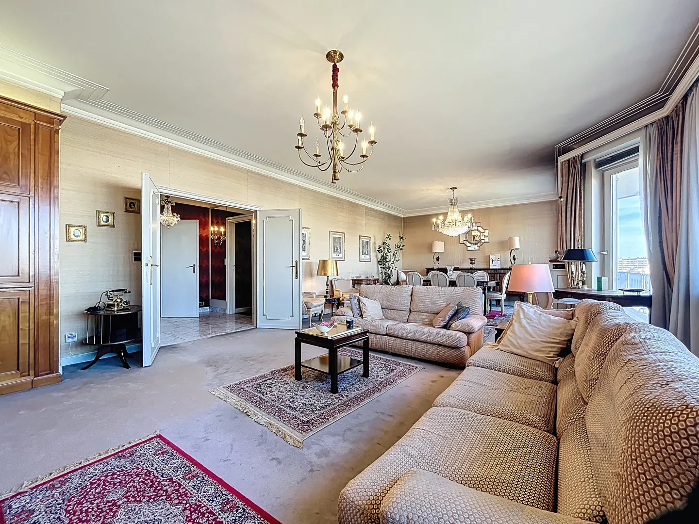 A wide shot of a living room with beige sofas, a chandelier, and a dining area in the background.