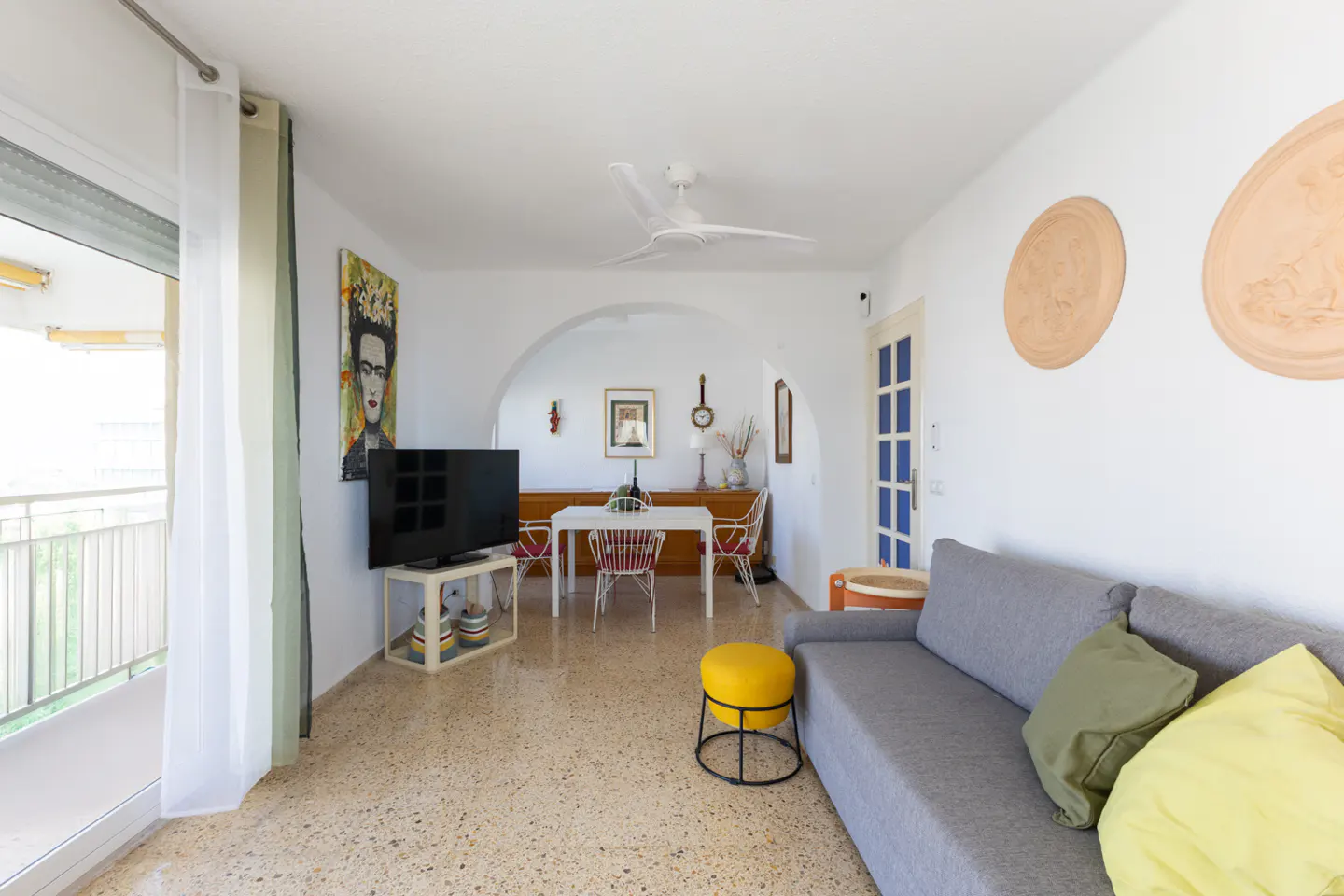 Bright living room with gray sofa, yellow pillows, and Frida Kahlo art. Dining area visible through an archway. Balcony with sheer curtains.