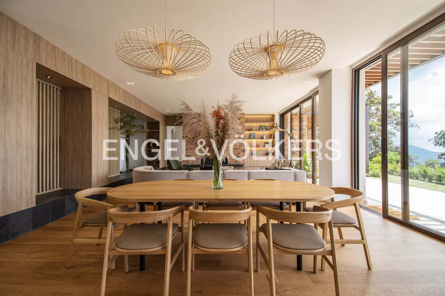A bright dining room features a wooden table with six chairs, two modern light fixtures, and a vase of flowers. Large windows offer a view of the green landscape.