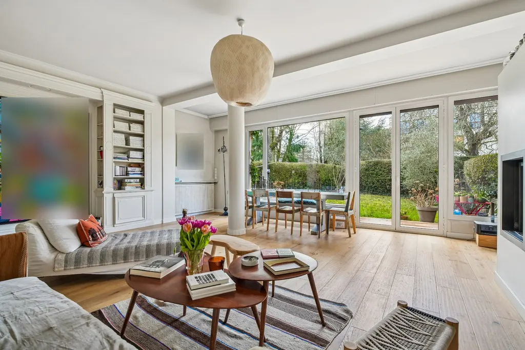 Bright living room with wood floors, sofas, and a dining table visible through large windows overlooking a green garden.