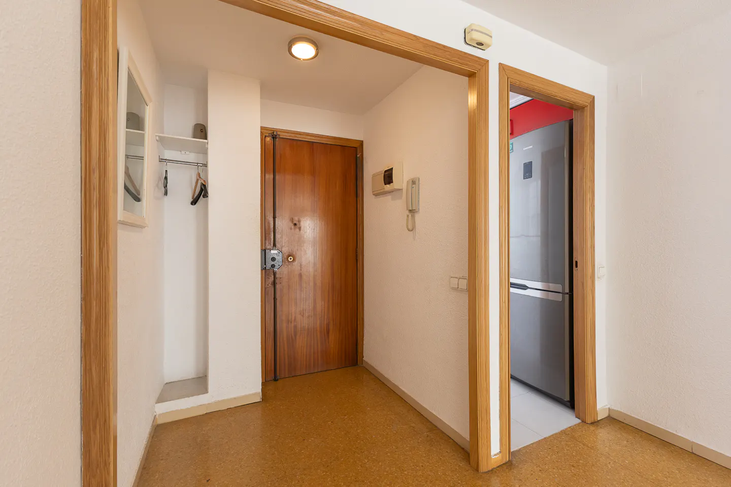 A hallway with a wooden front door, a coat rack, a wall-mounted intercom, and a doorway leading to a kitchen with a fridge.