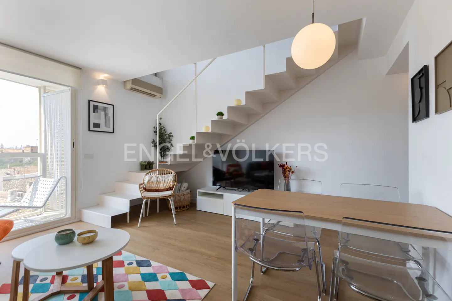 Bright, modern living room with white walls, wooden floors, and a staircase. A dining table with clear chairs sits near a TV. A colorful rug adds a pop of color.