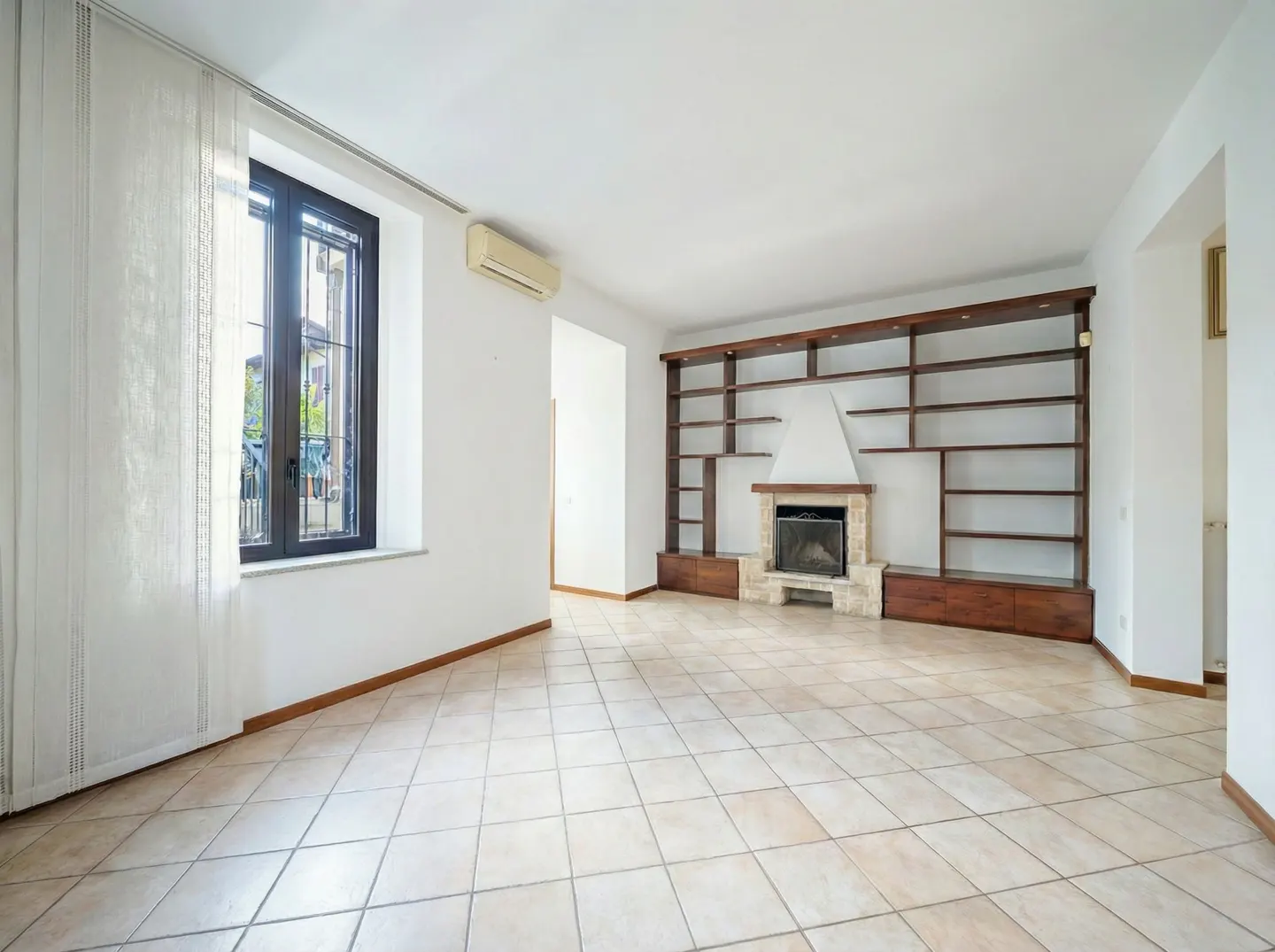 Bright, empty living room with tile floor, white walls, and a dark wood bookcase surrounding a stone fireplace. A window with sheer curtains is on the left.