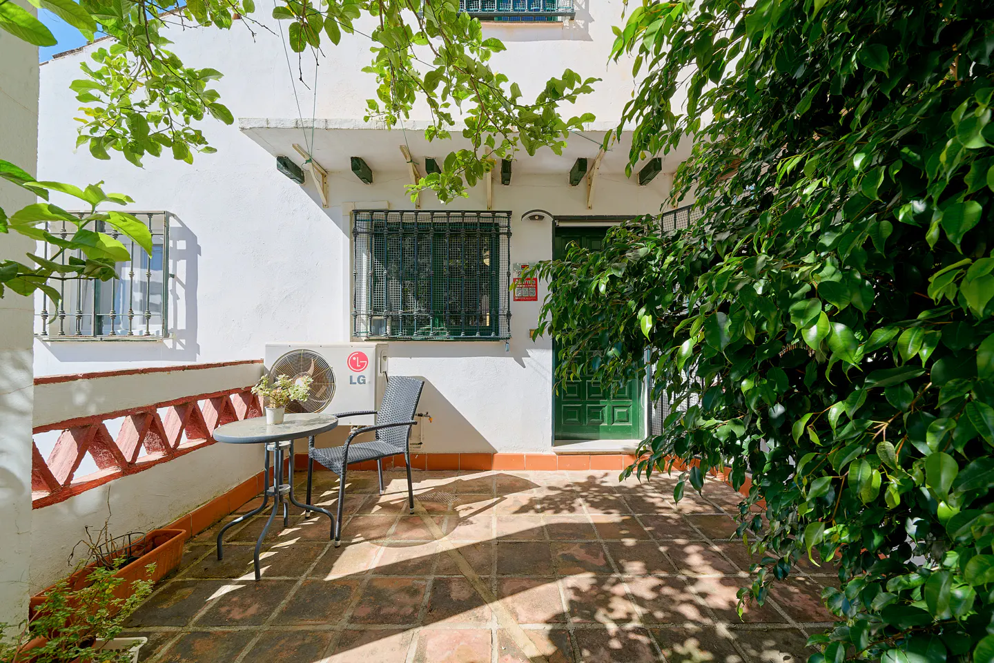 Exterior view of a white building with a green door and window bars. A table and chair sit on a brick patio, shaded by trees.