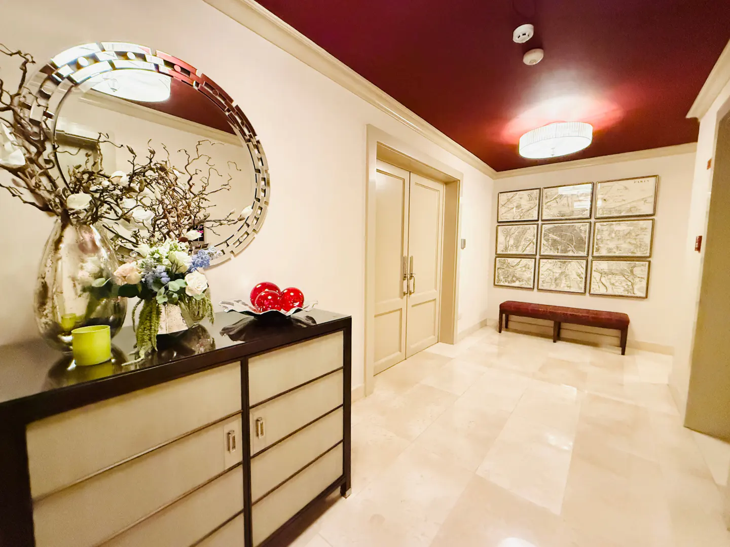 Elegant foyer with a burgundy ceiling, cream walls, and marble floors. A mirrored console table holds floral arrangements and red spheres. Artwork adorns the wall.
