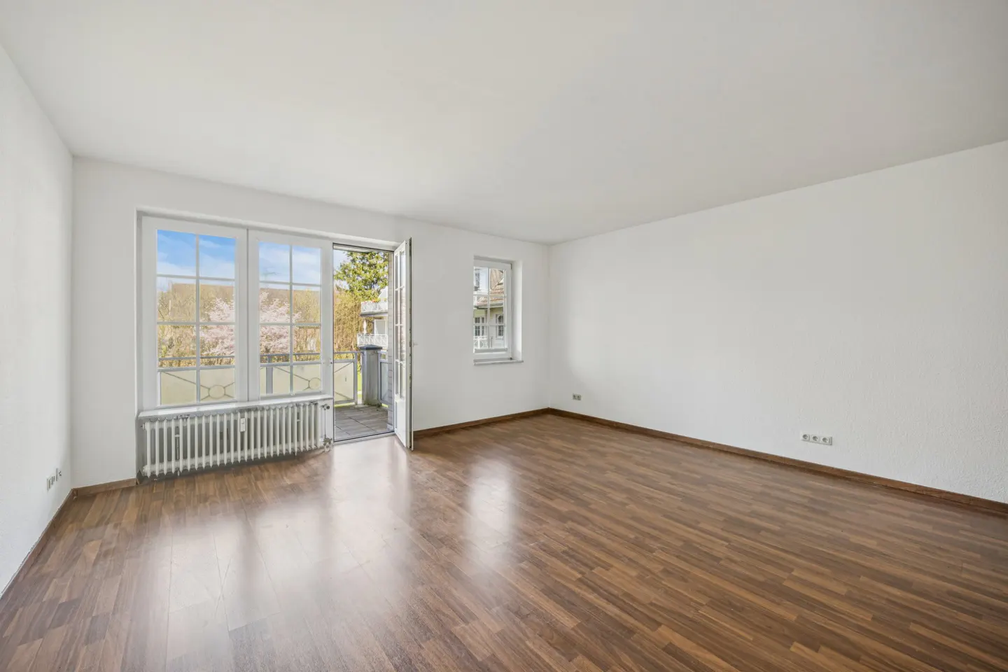 Empty room with wood floors, white walls, radiator, and open French doors to a balcony with a view of trees.