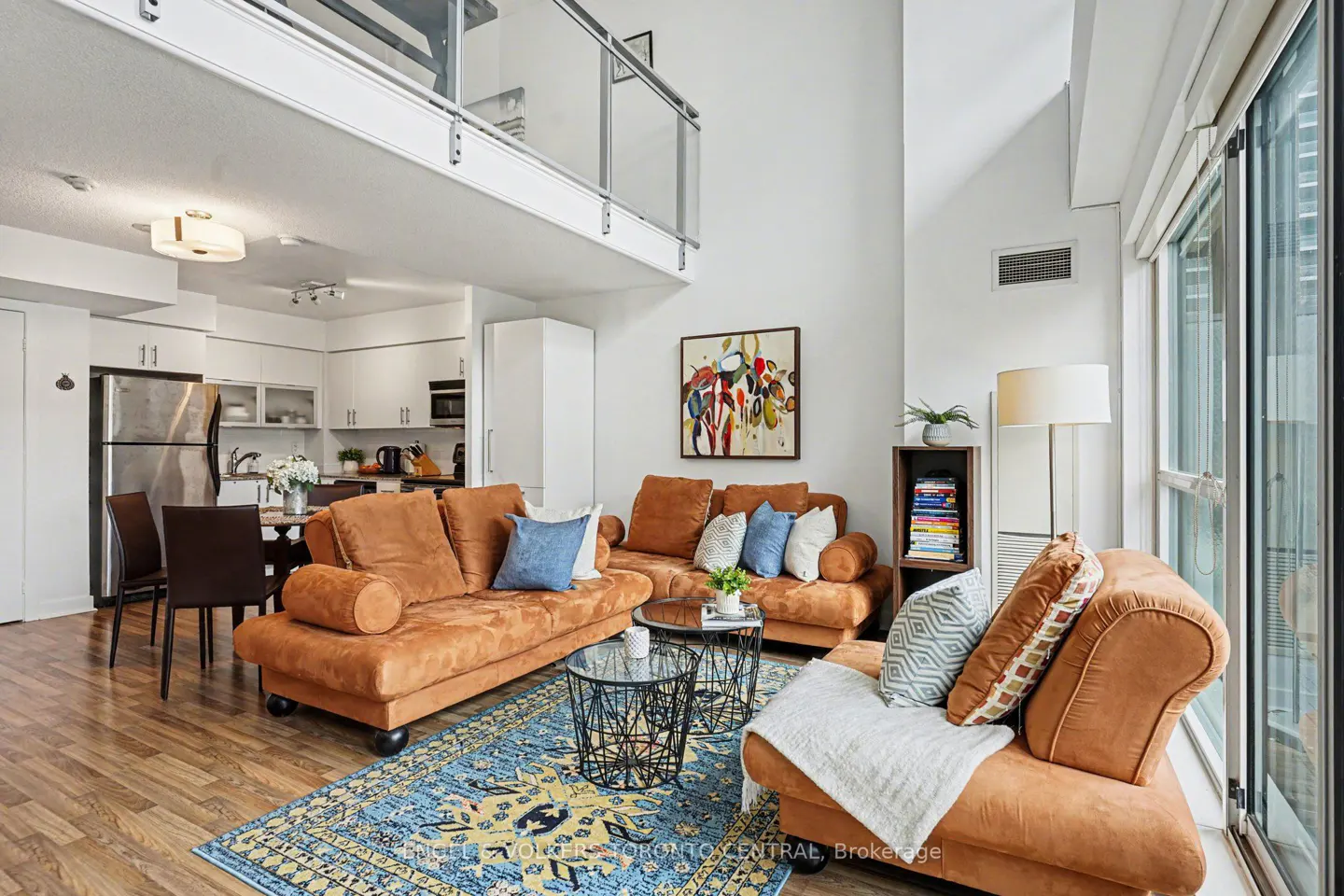 Bright, modern condo living room with brown sofas, blue rug, and loft above. Kitchen visible in the background.