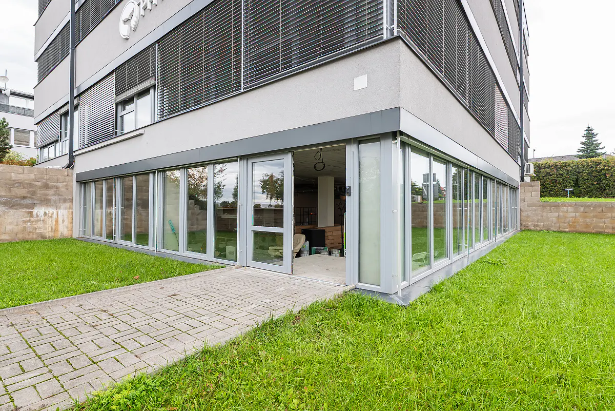 Exterior view of a modern gray building with a glass-enclosed ground floor, green lawn, and a stone-paved walkway leading to an open glass door.