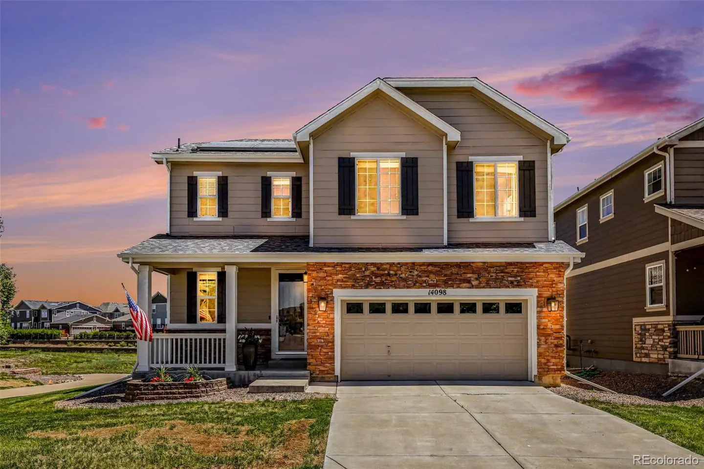 Two-story suburban home with tan siding, stone accents, and a two-car garage at sunset with a colorful sky.