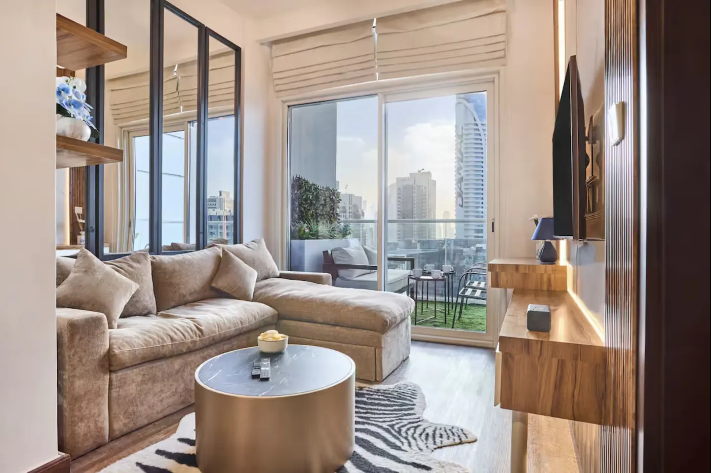 Living room with a beige sectional sofa, round coffee table, and zebra rug. Balcony with seating overlooks a city skyline.