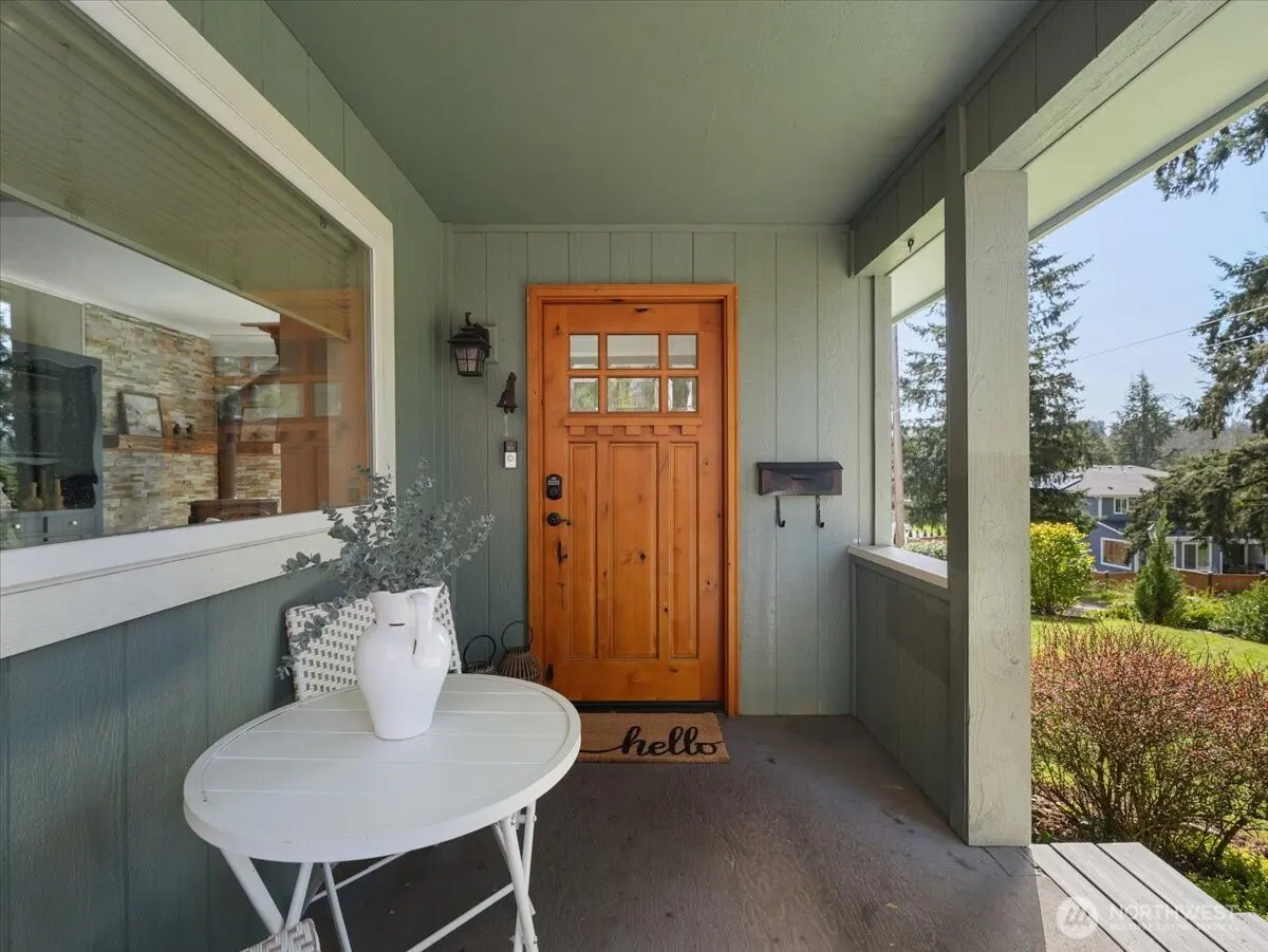 Covered porch with a wooden front door, a white table with a vase, and a view of a green lawn and trees.