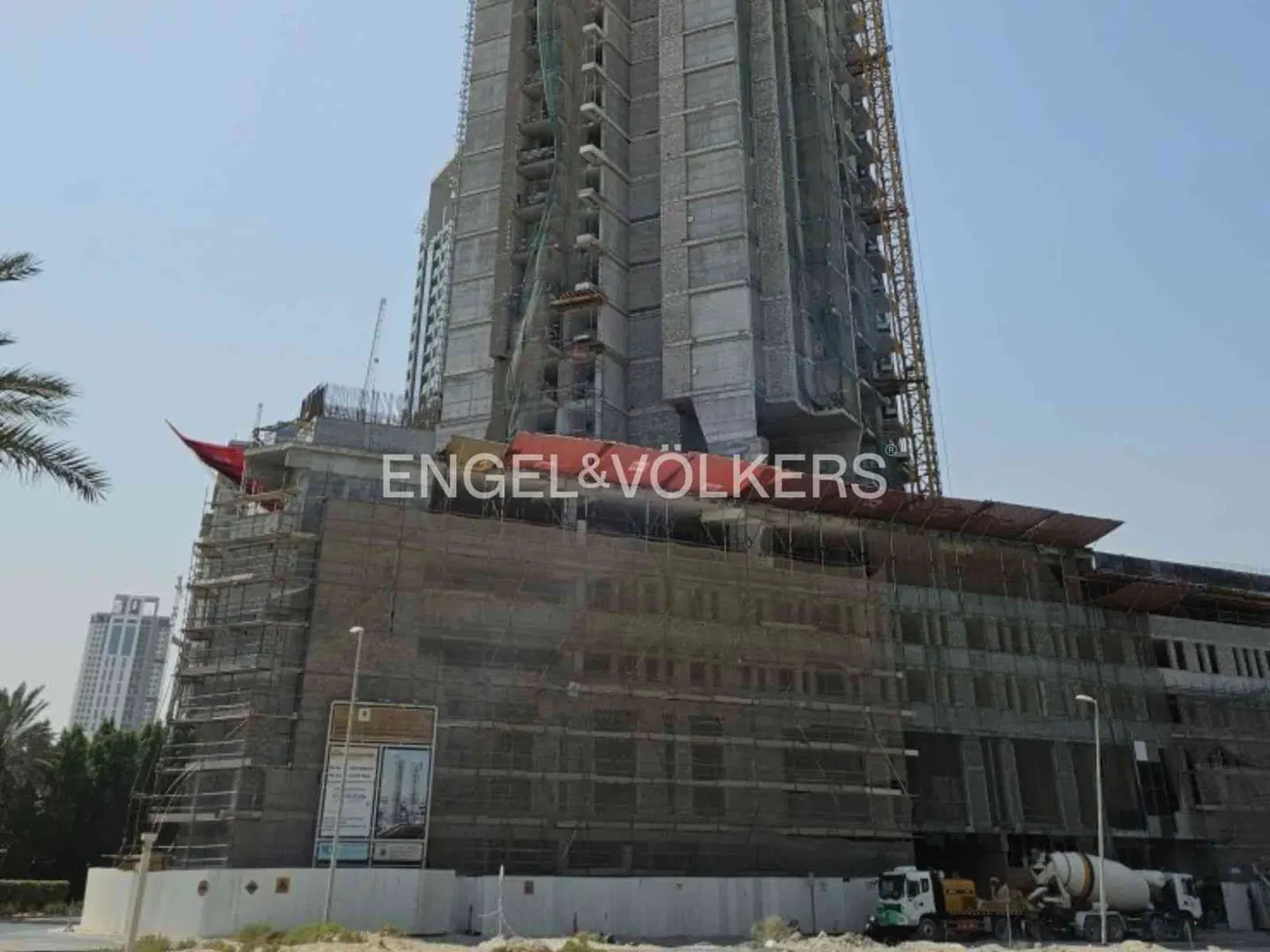 A tall building under construction, covered in scaffolding, with Engel & Völkers logo. Cement trucks are parked nearby.