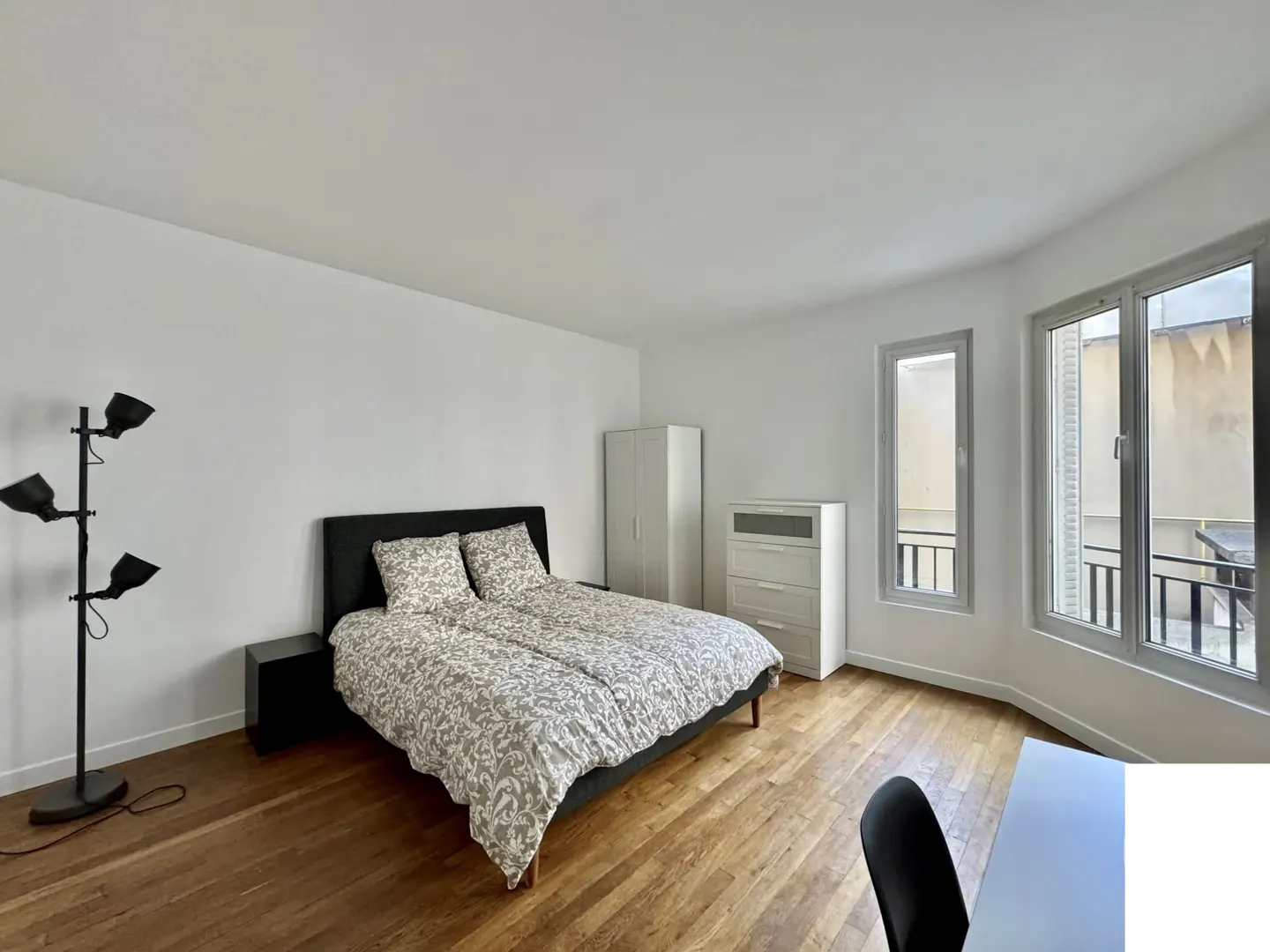 A bright bedroom with a bed, black headboard, patterned bedding, wood floors, and a black floor lamp. A white dresser and wardrobe are near windows.