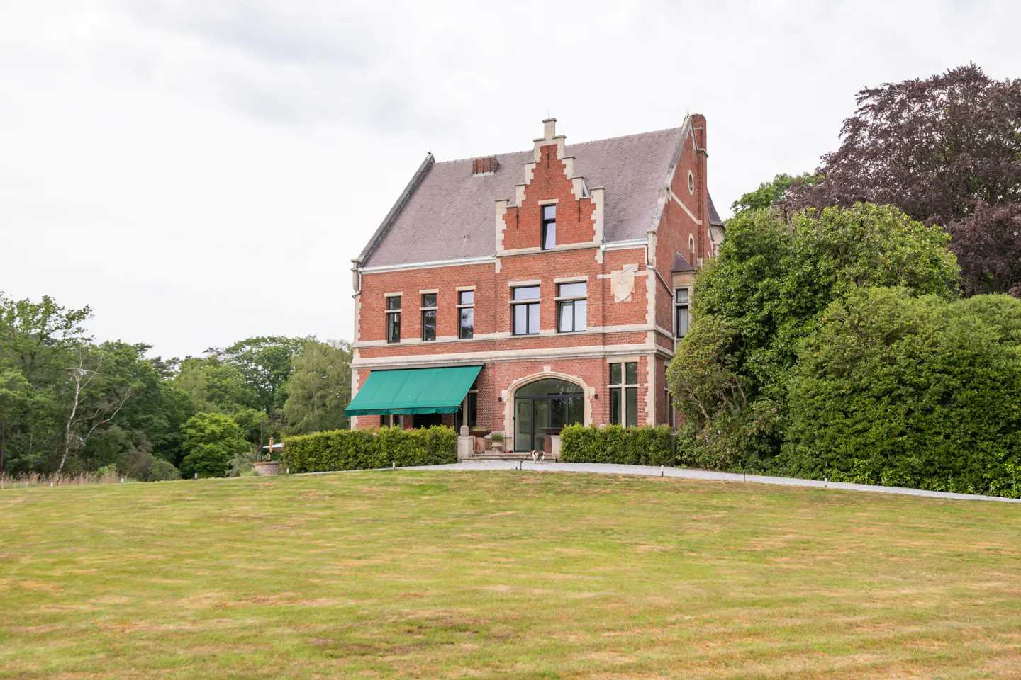 A large, three-story red brick mansion with a green awning, set behind a manicured lawn and surrounded by lush trees.