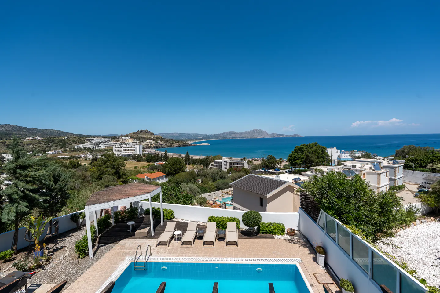 Aerial view of a blue swimming pool with lounge chairs, a pergola, and a view of the ocean and town in the background.