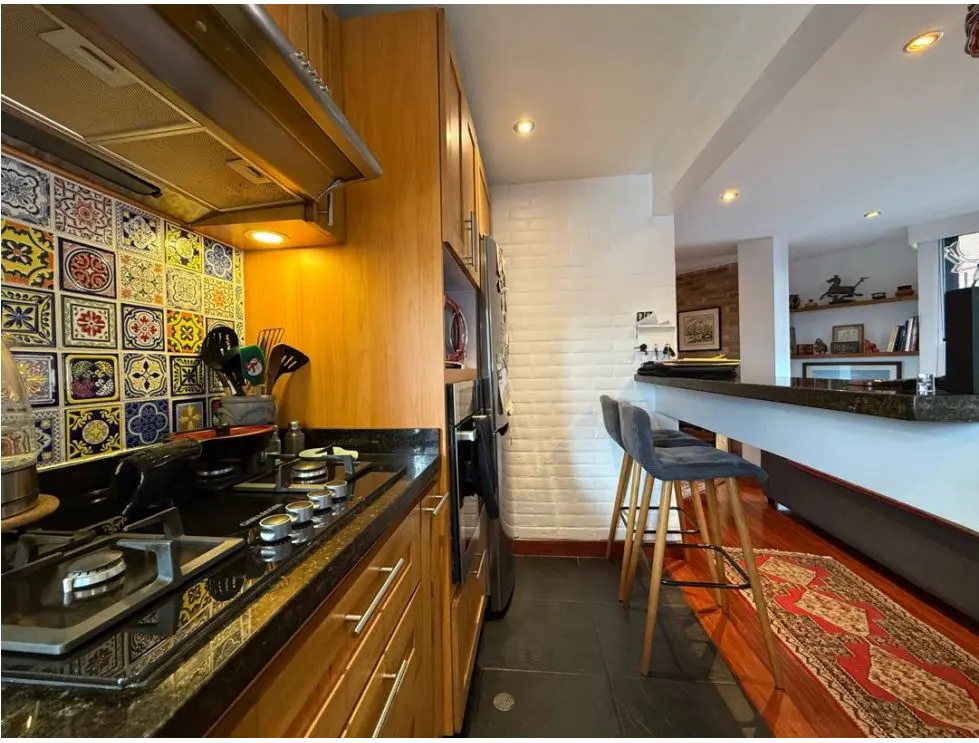 A kitchen with wood cabinets, black countertops, and colorful tile backsplash. A breakfast bar with two gray stools is visible.
