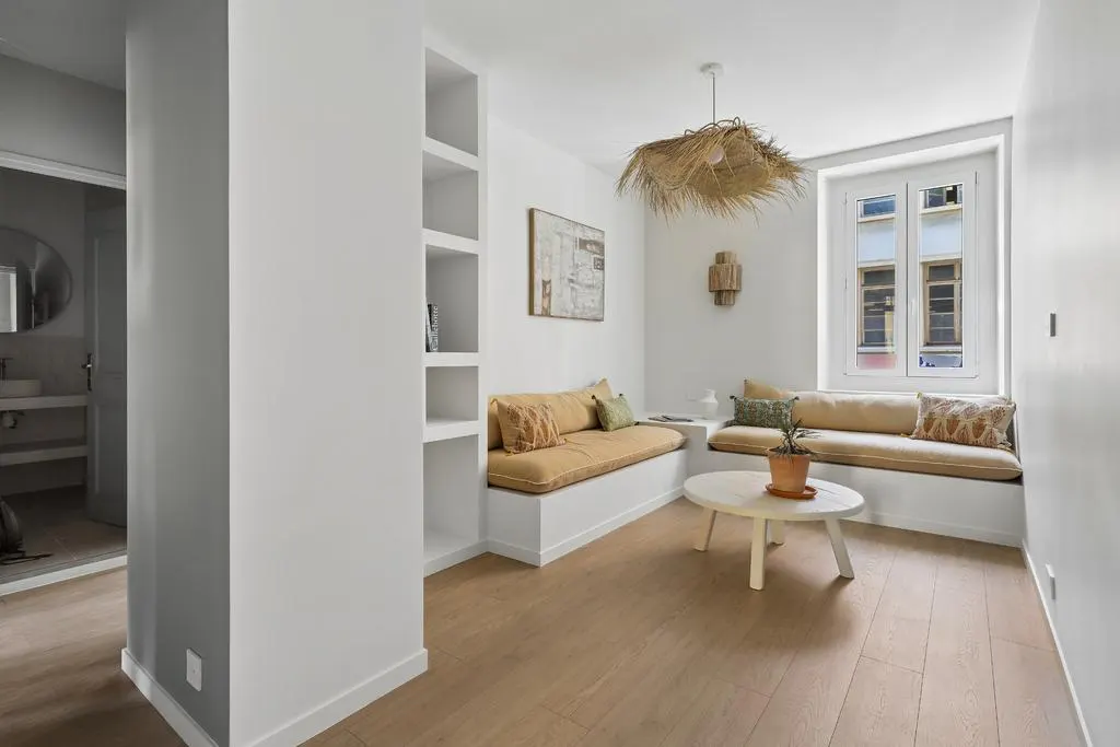 Bright living room with built-in seating, white walls, and wood floors. A round table sits in front of a window seat. A straw pendant light hangs above.