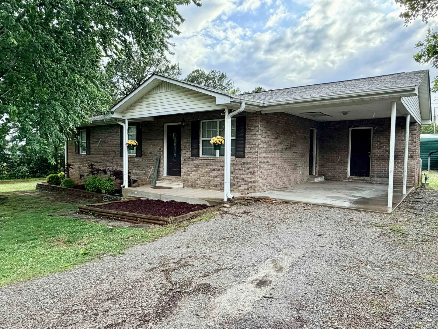 A single-story brick house with a carport and a gravel driveway. Yellow flowers hang from the windows.