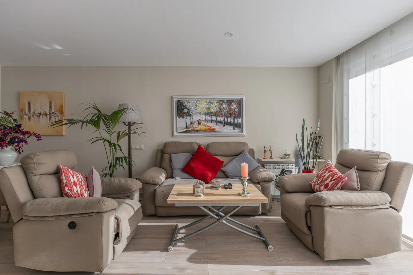 Living room with beige recliner chairs, sofa, and a light wood coffee table. Artwork hangs on the wall. A large window lets in natural light.