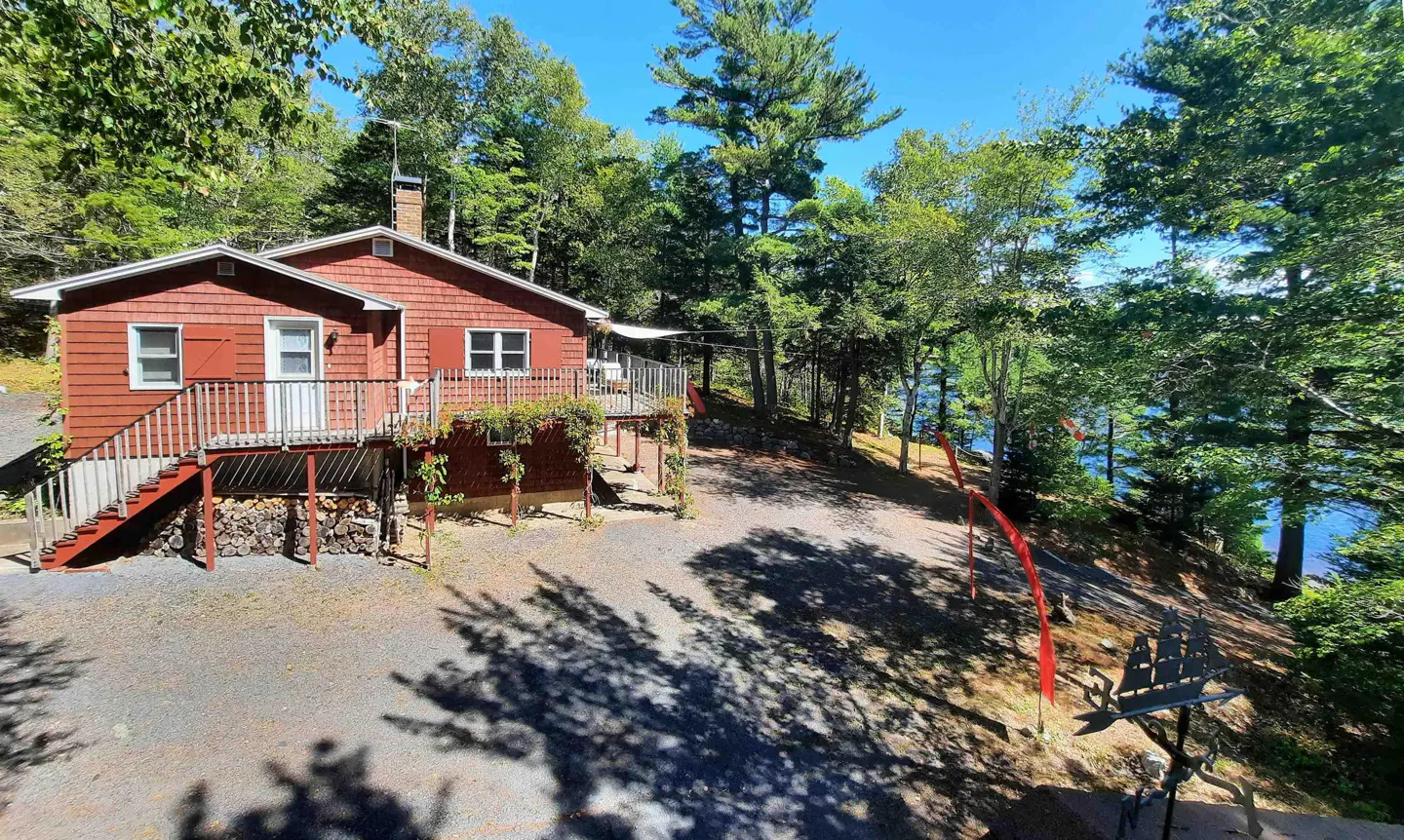 Exterior view of a red cabin with a deck, surrounded by trees and a glimpse of a lake.