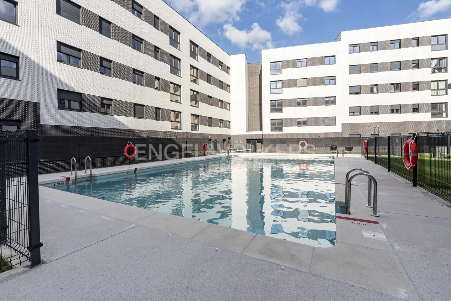 Apartment complex with a pool. The building is white and gray brick with dark windows. Red life preservers are on the fence.