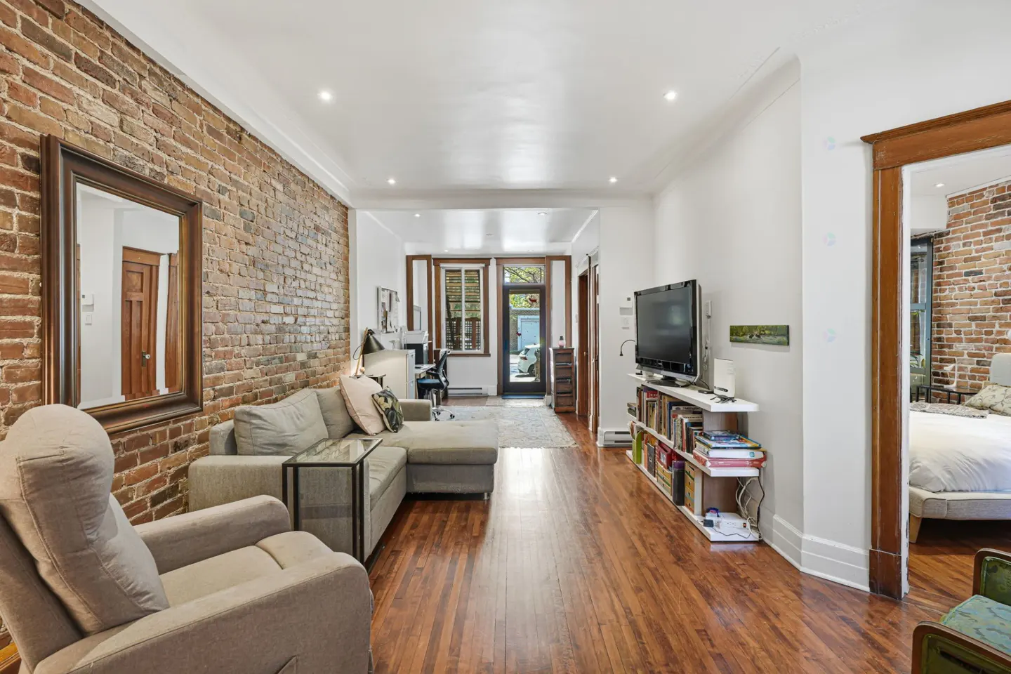 Living room with exposed brick wall, gray sofa, recliner, and hardwood floors. View to bedroom and office.