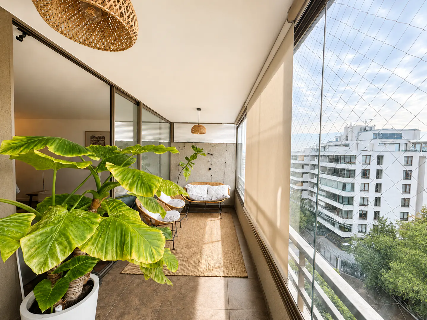 A sunlit balcony with wicker furniture, a jute rug, and a large potted plant. Outside, buildings and trees are visible through a safety net.