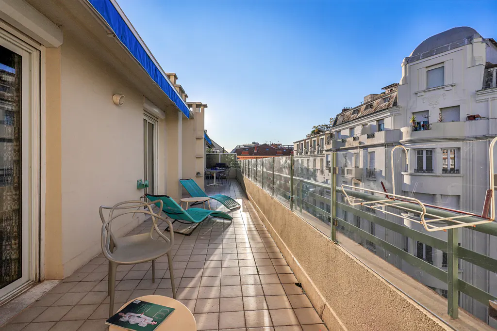 A sunny balcony with blue lounge chairs, a gray chair, and a small table with a magazine. A glass railing overlooks city buildings.