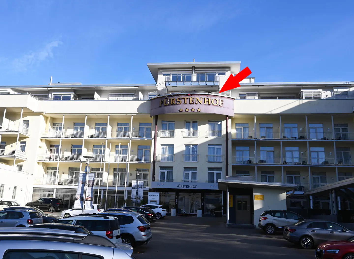 Exterior of the Furstenhof hotel, a multi-story building with balconies, under a blue sky. A parking lot with cars is in front.
