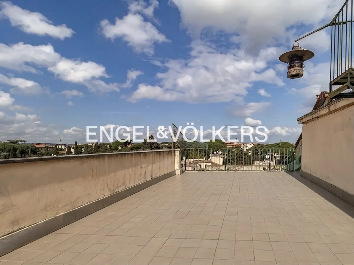 Wide rooftop view with beige tiles, low walls, and a metal railing overlooking trees and buildings under a blue, cloudy sky.
