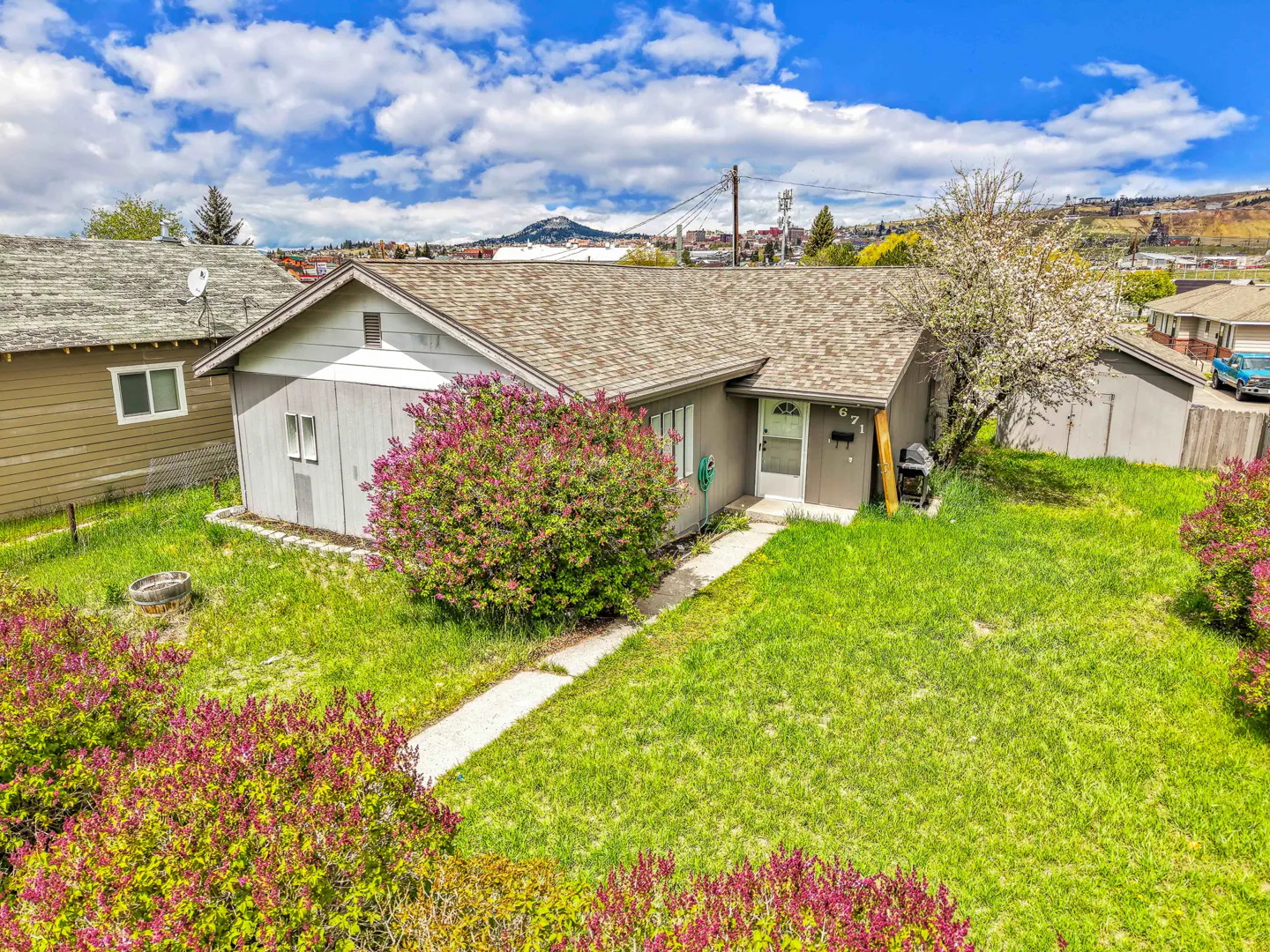 A single-story gray house with a brown roof, green lawn, and blooming bushes under a blue sky.