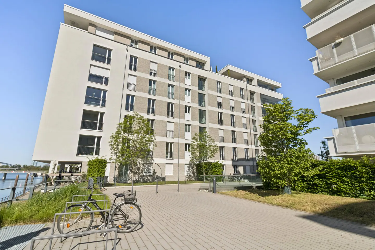 Modern apartment building with white and gray facade, green trees, and a bicycle parked in front on a sunny day.