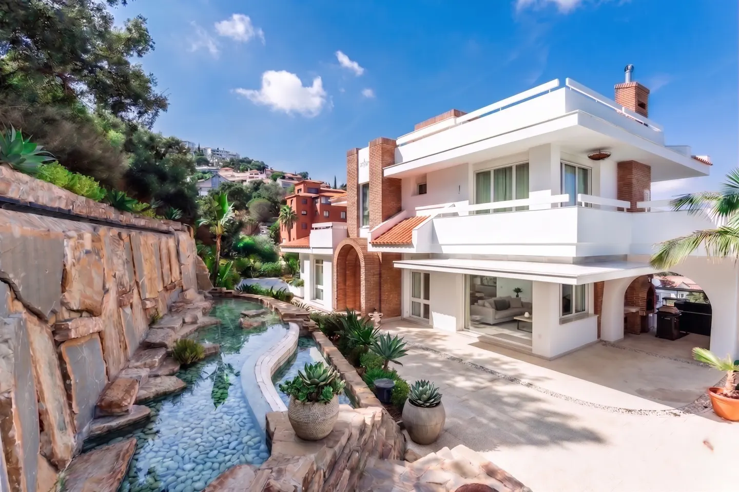 Exterior view of a modern white house with brick accents, a stone waterfall feature, and lush greenery under a blue sky.