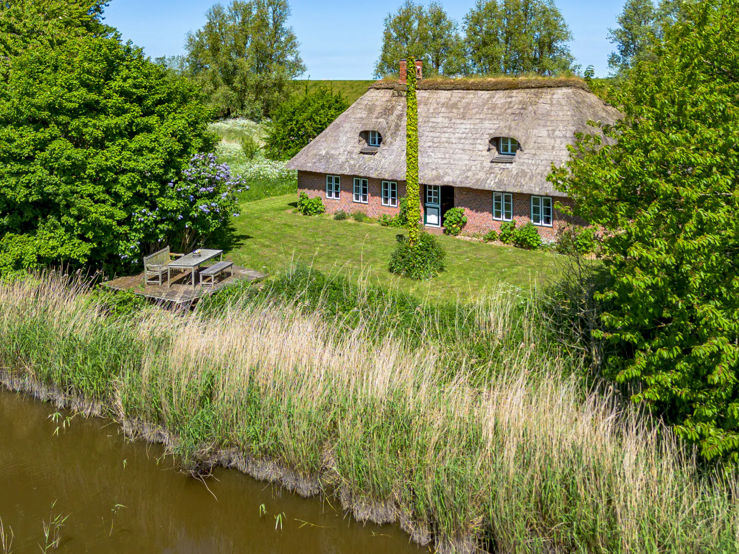 A charming red brick cottage with a thatched roof sits on a lush green lawn beside a canal with tall reeds.