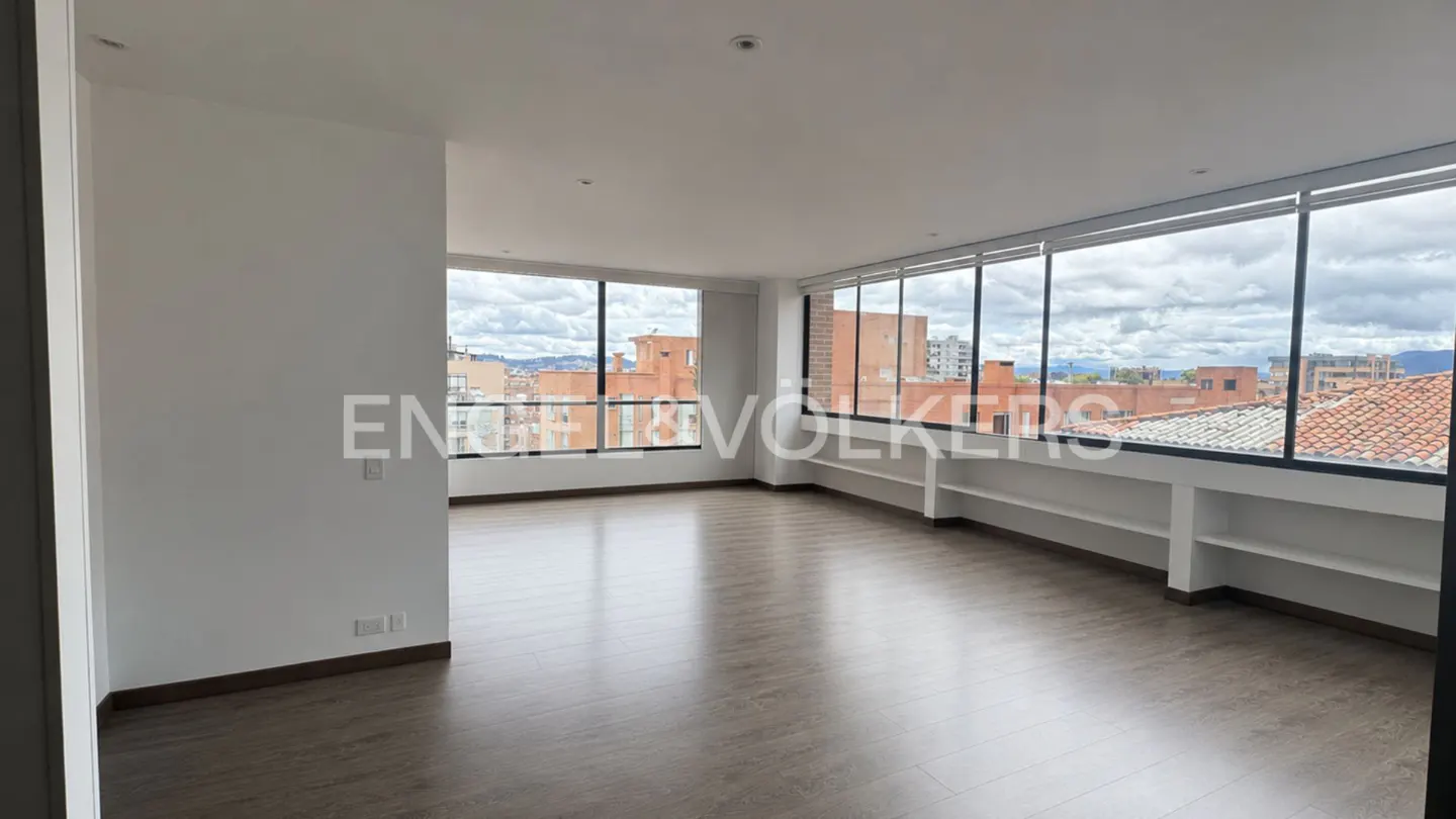 Empty modern room with light wood floors, white walls, built-in shelving, and large windows overlooking a city skyline.