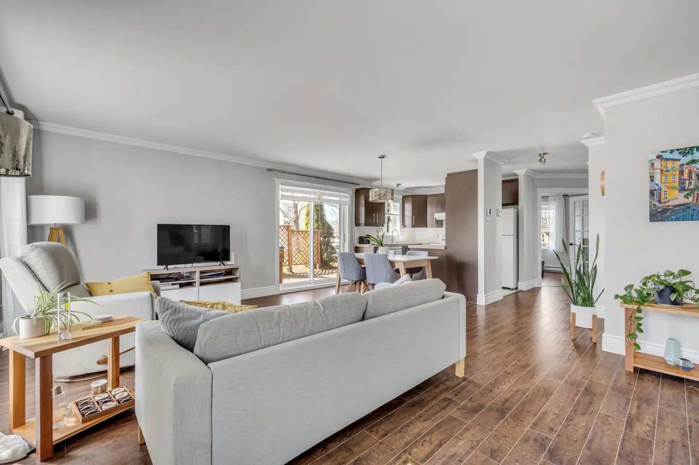 A bright, open-concept living room with hardwood floors, a gray sofa, TV, and a view into the kitchen and backyard.