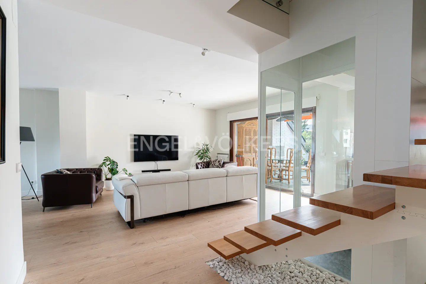 Bright, modern living room with white walls, light wood floors, and a white sofa. A staircase with wood treads and white risers is in the foreground.