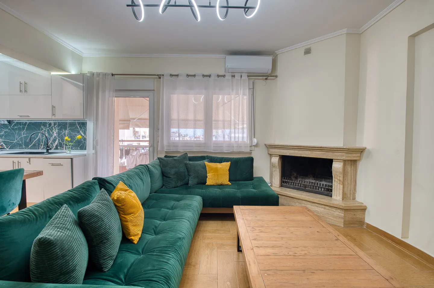 Living room with a green sectional sofa, yellow pillows, a wooden coffee table, and a stone fireplace. Kitchen in the background.