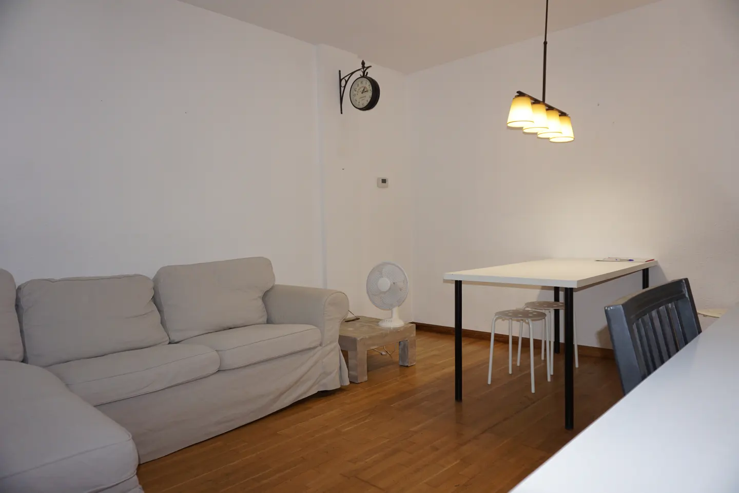 A bright living room with a gray sofa, white walls, and a wooden floor. A white table with stools sits under a pendant light. A wall clock and fan are also visible.
