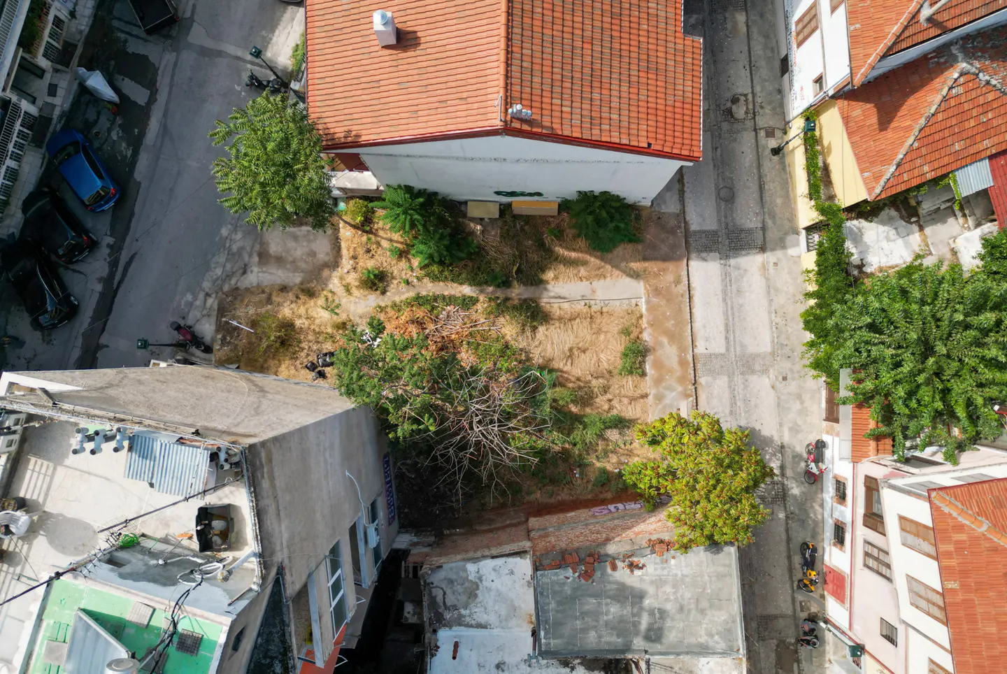 Aerial view of a vacant lot with dry grass, surrounded by buildings with red tile roofs and narrow streets.