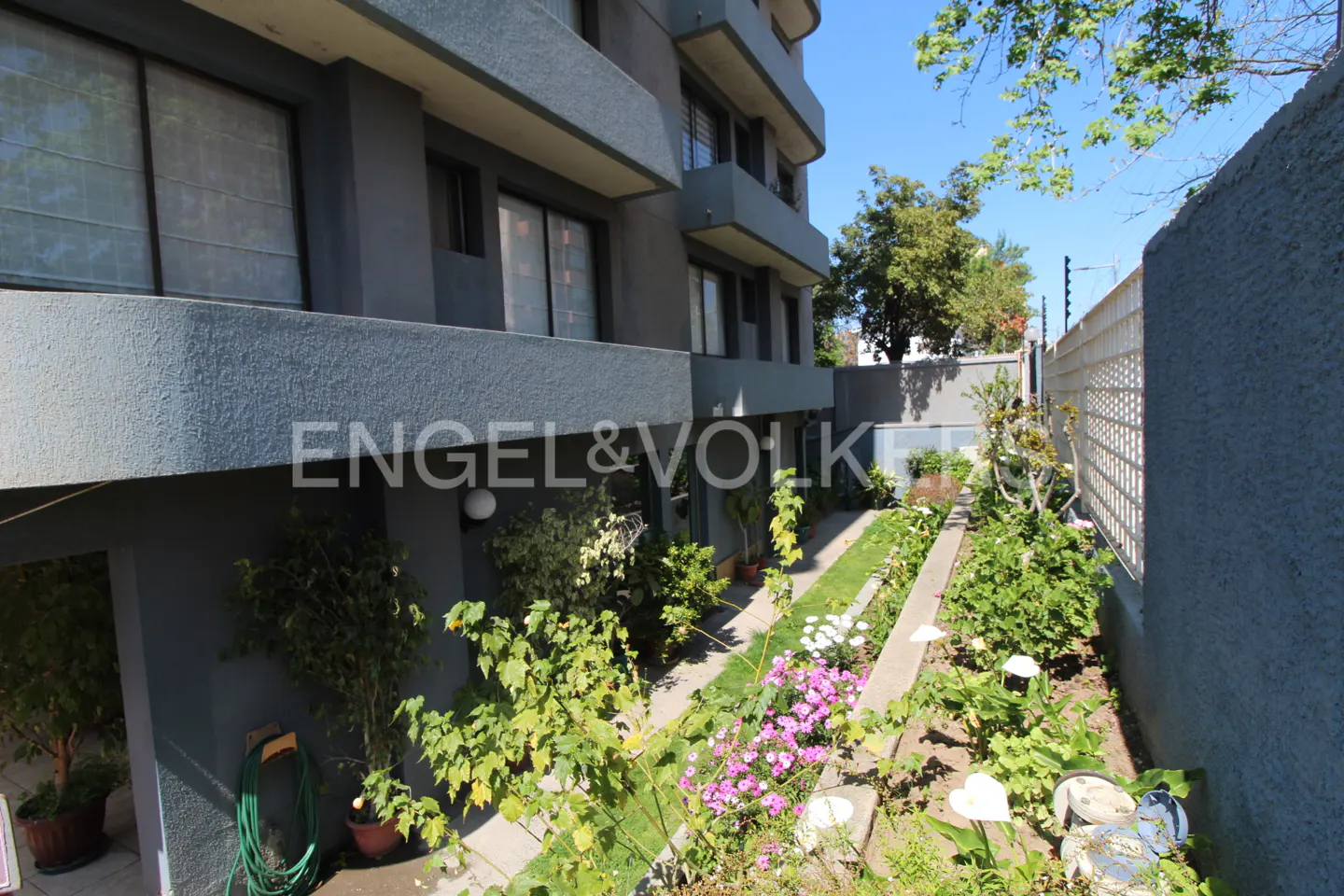Exterior of a gray apartment building with a garden, flowers, and the Engel & Volkers logo.